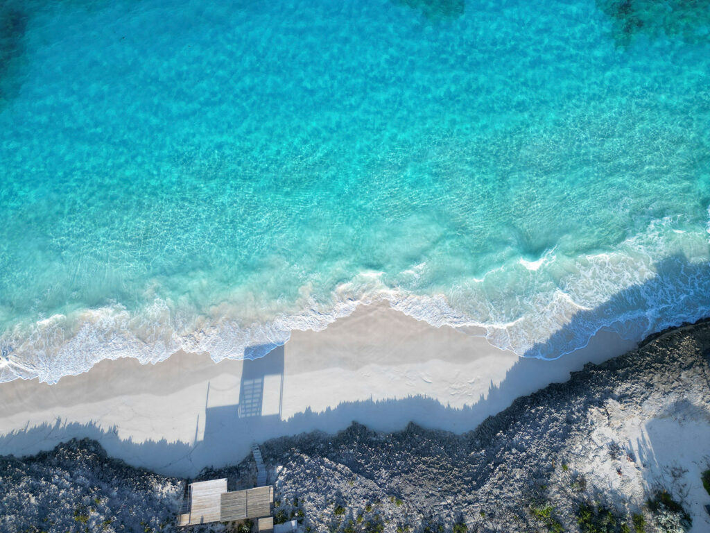 Aerial view of the beach in front of Indigo House Exuma, showing turquoise waves meeting white sand.