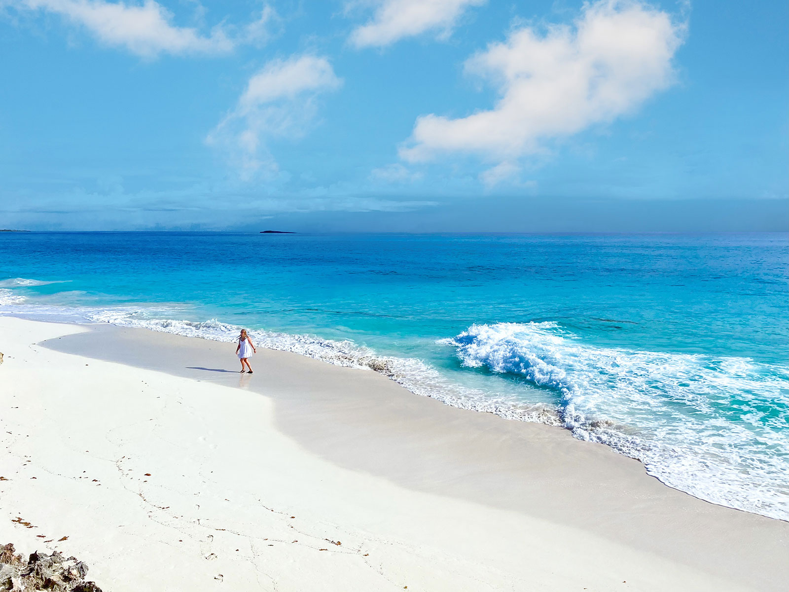 Indigo-House_beach-2_1600 Woman walking along a pristine white sand beach in Exuma with turquoise waves and a distant cay on the horizon.