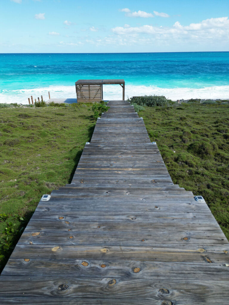View from Indigo House Exuma of a private wooden boardwalk leading through greenery to the beach and turquoise sea.