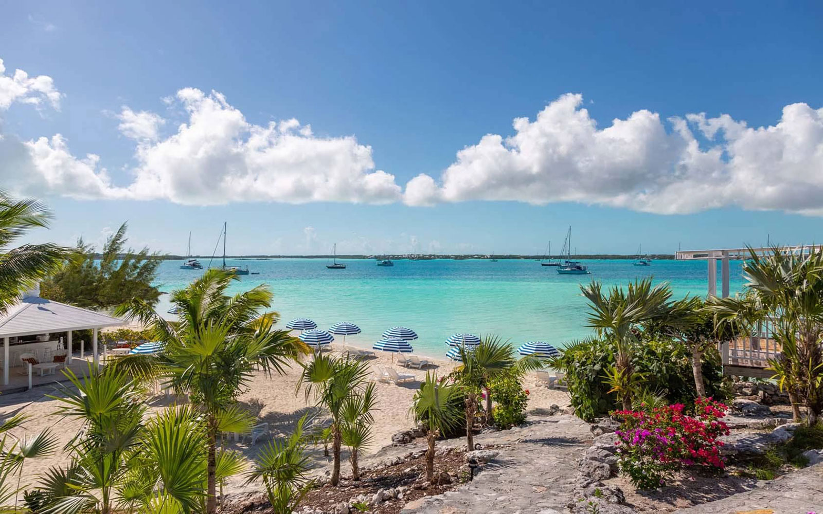 Indigo-House_peace-and-plenty-1_1600 View of the beach at Peace and Plenty Resort in Exuma, with striped blue umbrellas, lounge chairs, and turquoise water with anchored sailboats.