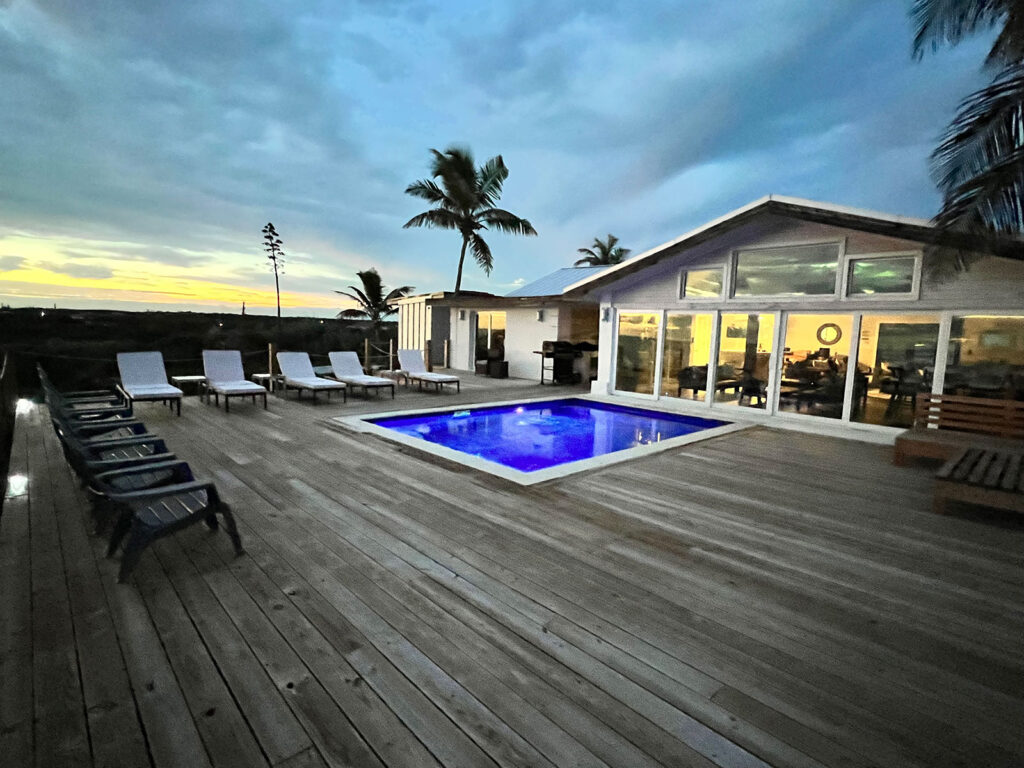 Evening view of the lit pool and spacious wooden deck at Indigo House Exuma with palm trees and sunset sky.