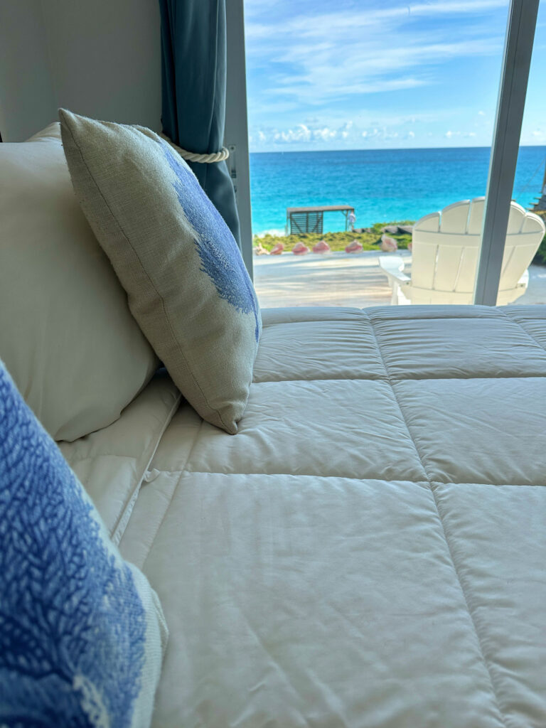 Close-up of a cozy bed with soft blue accents and ocean views through sliding glass doors.