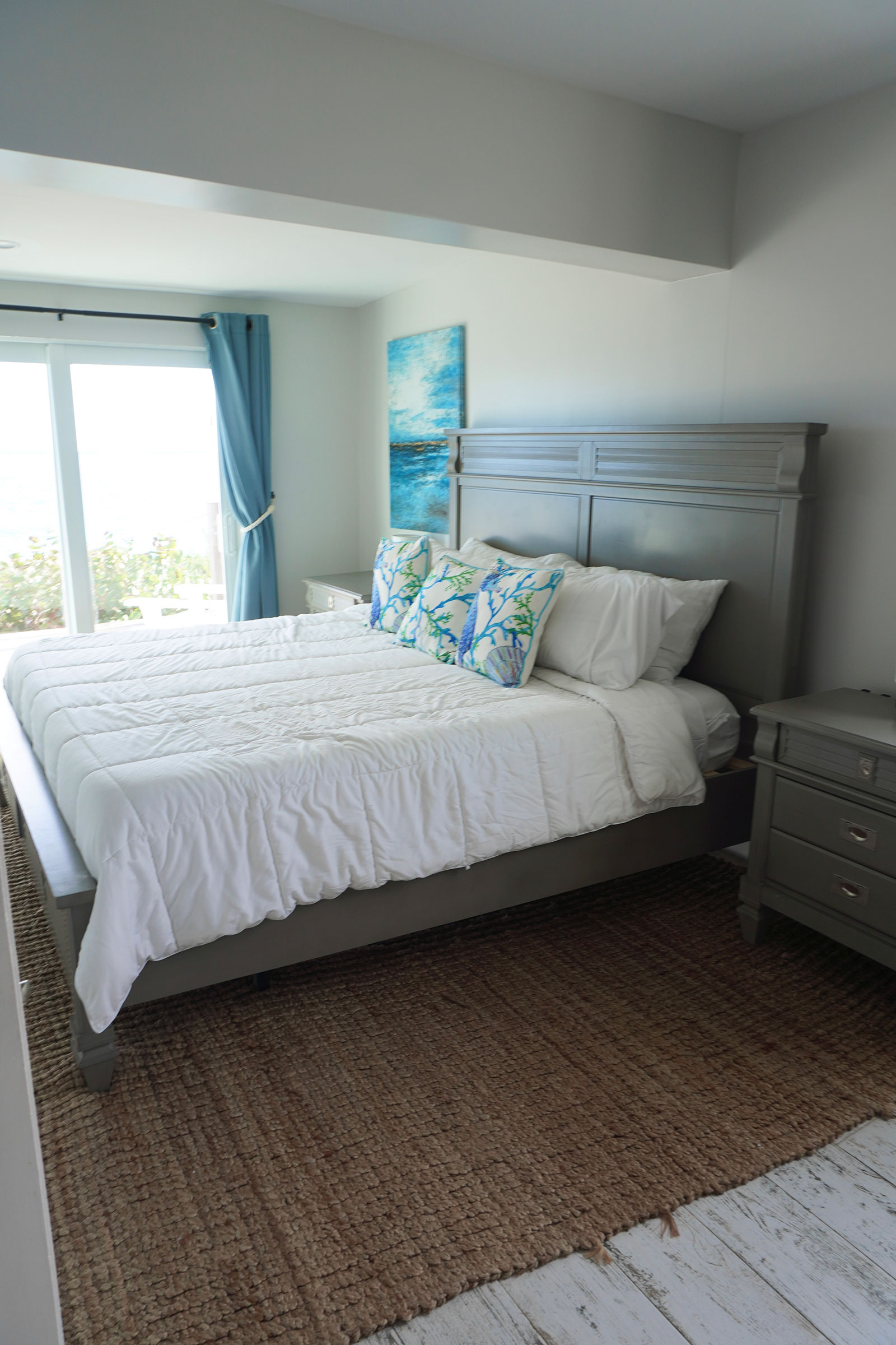 Indigo-House_suites-2_1600 Cozy bedroom at Indigo House Exuma with a gray wood bed frame, coral-themed pillows, and ocean views through sliding glass doors.