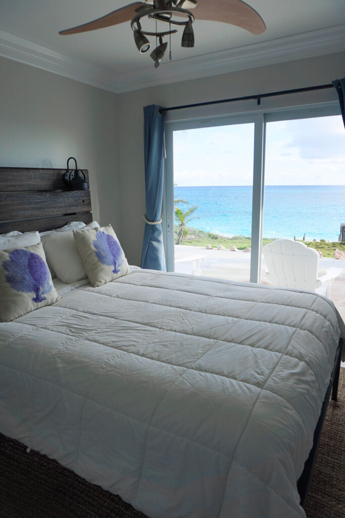 Oceanfront bedroom at Indigo House Exuma with light bedding, purple pillows, and a view of turquoise water.