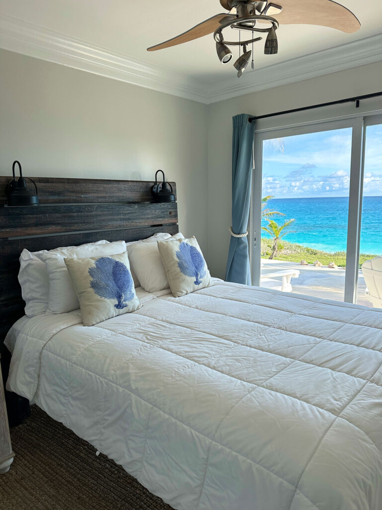 Bedroom suite at Indigo House Exuma with wood headboard, coastal pillows, and ocean views through glass doors.